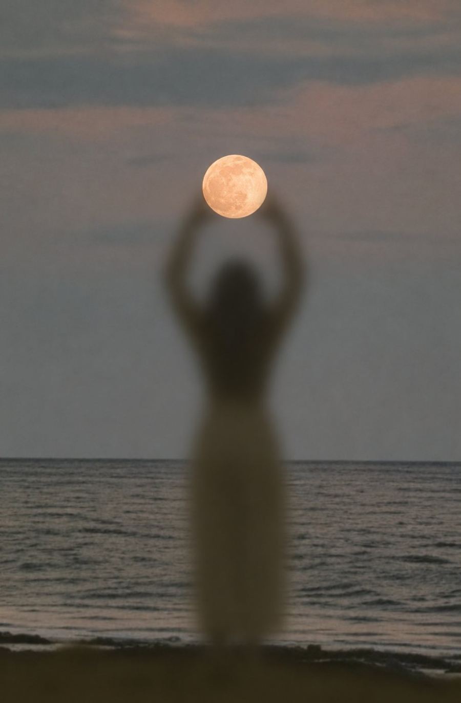 Silueta de una mujer junto al mar sosteniendo la luna llena entre las manos